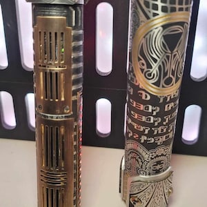 May include: Two silver and black lightsaber hilts with intricate engravings. One hilt has a brass-colored section with a grid pattern and red lights. The other hilt has a gold-colored circle with a design inside and text in a foreign language.