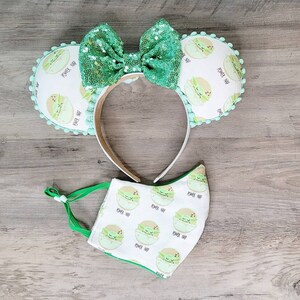 K&ouml;nnte beinhalten: Ein gr&uuml;nes und wei&szlig;es Stirnband mit einer gr&uuml;nen Schleife und einem Muster von schlafenden Baby Yoda-Charakteren. Das Stirnband wird mit einer gr&uuml;nen und wei&szlig;en Gesichtsmaske mit dem gleichen Muster kombiniert.