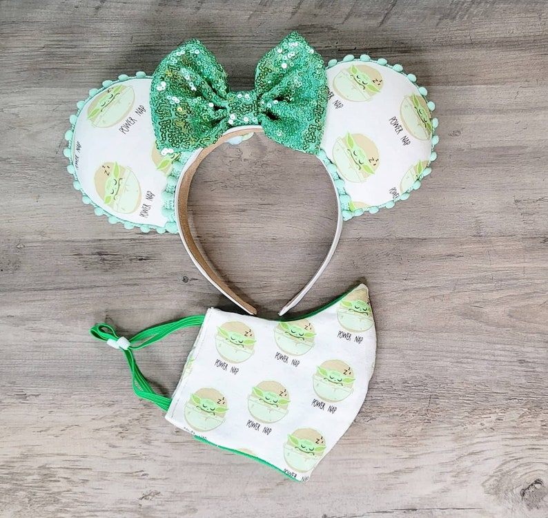K&ouml;nnte beinhalten: Ein gr&uuml;nes und wei&szlig;es Stirnband mit einer gr&uuml;nen Schleife und einem Muster von schlafenden Baby Yoda-Charakteren. Das Stirnband wird mit einer gr&uuml;nen und wei&szlig;en Gesichtsmaske mit dem gleichen Muster kombiniert.