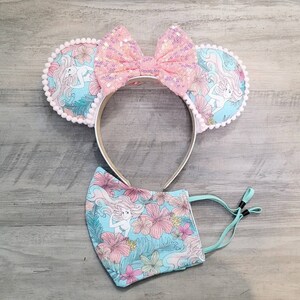 Pode incluir: Um par de orelhas de rato rosa e azul com um la&ccedil;o rosa e uma estampa de sereia. As orelhas s&atilde;o presas a uma faixa de cabe&ccedil;a. Tamb&eacute;m est&aacute; inclu&iacute;da uma m&aacute;scara facial combinando azul e rosa com uma estampa de sereia.
