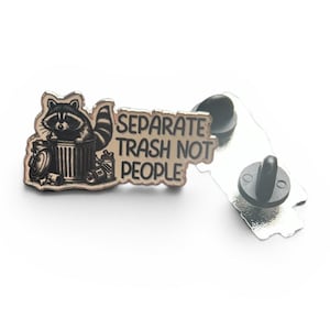 Könnte beinhalten: Ein silberner Metall-Ansteckpin mit einem Waschbären, der aus einem Mülleimer schaut. Der Pin trägt den Text "Separate Trash Not People". Der Pin ist schwarz und silber.