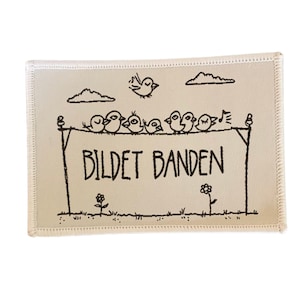 Könnte beinhalten: Rechteckiger Aufnäher in Cremefarbe mit schwarzem Zierstich. Der Aufnäher zeigt eine skurrile Illustration von Vögeln, die auf einer Wäscheleine sitzen, mit dem Text "BILDE BANDEN" darunter. Zwei kleine Blumen und Wolken sind ebenfalls vorhanden.