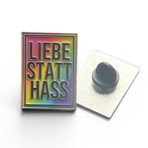 Könnte beinhalten: Rechteckiger Emaille-Pin mit den Worten "LIEBE STATT HASS" in schwarzen Blockbuchstaben, vor einem Regenbogen-Farbverlauf. Der Pin hat einen silbernen Rand und einen schwarzen Pin-Rücken. Der Pin ist ein Statement-Stück.