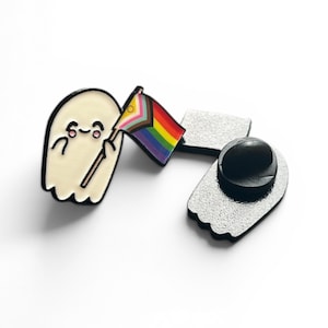 Könnte beinhalten: Drei Emaille-Anstecknadeln: ein lächelnder Geist, der eine Regenbogen-Pride-Flagge hält, und die Rückseiten der Geister- und Flaggen-Anstecknadeln. Die Anstecknadeln haben schwarze Umrisse und befinden sich auf weißem Hintergrund. Der Geist ist cremefarben.