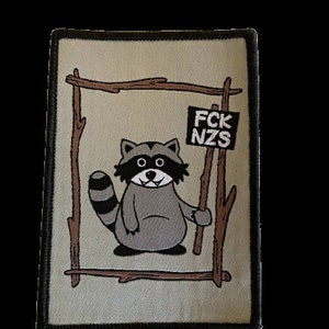 Könnte beinhalten: Ein grauer Waschbär mit einer schwarzen Maske und einem Schwanz steht in einem Holzrahmen. Der Waschbär hält einen Stock mit einem Schild, auf dem "FCK NZS" steht.