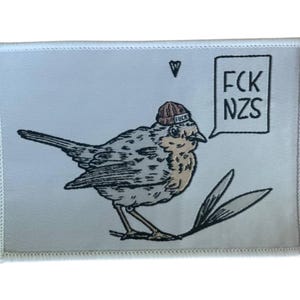 Könnte beinhalten: Ein weißes Patch mit einer Vogelillustration, die eine braune Mütze trägt. Der Vogel befindet sich vor einer Sprechblase mit dem Text "FCK NZS". Das Patch hat einen weißen Rand und ein kleines schwarzes Blatt.