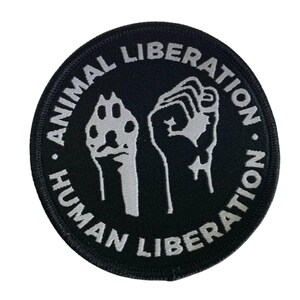 Könnte beinhalten: Ein runder, schwarzer Aufnäher mit weißem Text und Bildern. Der Text lautet "ANIMAL LIBERATION" und "HUMAN LIBERATION". Das Bild zeigt einen Pfotenabdruck und eine Faust. Der Aufnäher ist für Kleidung oder Accessoires.