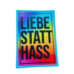 Könnte beinhalten: Ein regenbogenfarbener Patch mit dem Text "LIEBE STATT HASS" in Schwarz.