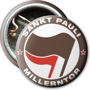 Könnte beinhalten: Ein runder, brauner Button mit dem weißen Schriftzug "Sankt Pauli Millerntor". Der Button zeigt ein braunes Flaggen-Design mit einem roten Streifen und zwei roten Sternen. Er hat eine Metallnadel auf der Rückseite.