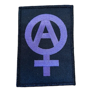 Könnte beinhalten: Ein rechteckiger bestickter Aufnäher mit schwarzem Hintergrund und einem mittig platzierten violetten Symbol. Das Design besteht aus einem großen A in einem Kreis, mit einem Kreuz am unteren Ende, das ein kombiniertes Anarchie- und Frauensymbol bildet.