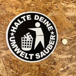 Könnte beinhalten: Ein runder Aufkleber in Schwarzweiß mit dem Text "Halte Deine Umwelt Sauber" und einer Strichmännchenfigur, die Müll in einen Mülleimer mit einem Hakenkreuz wirft.