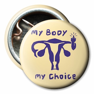Könnte beinhalten: Ein gelber Knopf mit einer lila Gebärmutter und einer Hand mit erhobenem Mittelfinger. Der Text auf dem Knopf lautet "My Body My Choice".