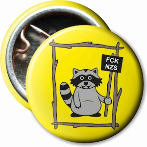 Könnte beinhalten: Ein gelber Knopf mit einem Cartoon-Waschbär, der ein Schild mit der Aufschrift "FCK NZS" hält. Der Waschbär befindet sich in einem Holzrahmen.