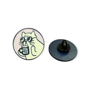Könnte beinhalten: Runder Emaille-Anstecker mit einer Cartoon-Katze, die eine Sonnenbrille trägt und eine Tasse hält. Auf der Tasse steht "FCK NZS". Die Katze zeigt den Mittelfinger. Der Anstecker hat einen hellrosa Hintergrund und eine dunkelblaue Rückseite.