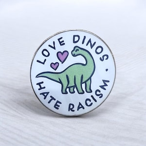 Könnte beinhalten: Ein runder Emaille-Pin mit weißem Hintergrund, der eine hellgrüne Illustration eines langhalsigen Dinosauriers, zwei kleine violette Herzen und den schwarzen Text LOVE DINOS HATE RACISM in kreisförmiger Anordnung zeigt.