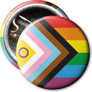 Könnte beinhalten: Zwei runde Ansteckbuttons. Ein Button zeigt ein Regenbogen-Design, während der andere eine progressive Pride-Flagge mit gelbem Hintergrund und einem lila Kreis zeigt. Die Buttons haben ein glänzendes Finish.