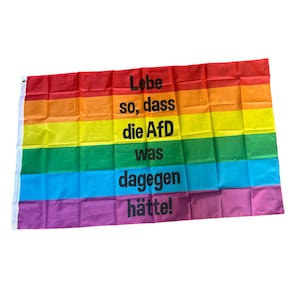 Könnte beinhalten: Eine Regenbogenflagge mit dem deutschen Text "Lebe so, dass die AfD was dagegen hätte!" in Schwarz. Die Flagge hat horizontale Streifen in Rot, Orange, Gelb, Grün, Blau und Lila.