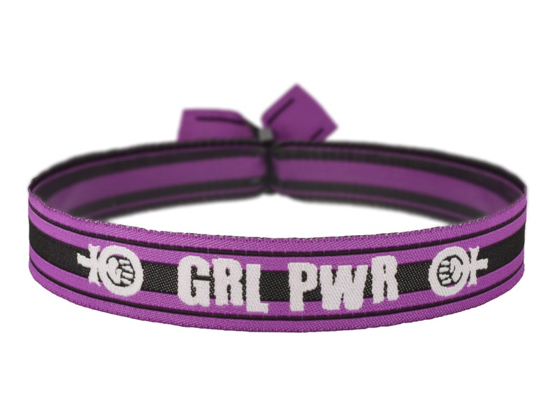 May include: A purple and black elastic hair tie with the text "GRL PWR" in white. The hair tie features a repeating pattern of a clenched fist and a female symbol. The tie is tied in a knot.