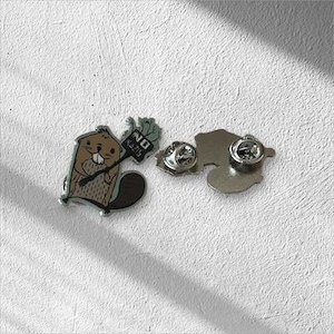 Könnte beinhalten: Ein Metall-Ansteckpin mit einem Cartoon-Biber, der ein Schild mit der Aufschrift "NO NAZIS" hält. Der Biber ist braun und beige mit einem dunkelbraunen Schwanz. Der Pin hat einen silbernen Rand und einen Butterfly-Verschluss.
