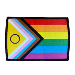 Könnte beinhalten: Rechteckiger Aufnäher mit Regenbogenflaggen-Design und schwarzem Rand. Die Flagge enthält die traditionellen Regenbogenfarben, einen gelben Bereich mit einem lila Kreis und einen Bereich mit rosa, hellblauen und braunen Dreiecken.