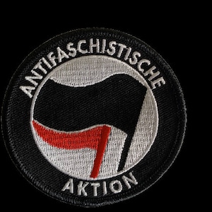 Könnte beinhalten: Ein gestickter schwarzer und weißer Aufnäher mit dem Text "ANTIFASCHISTISCHE AKTION" in einem Kreis. Der Aufnäher zeigt eine schwarze Faust mit einem roten Streifen.