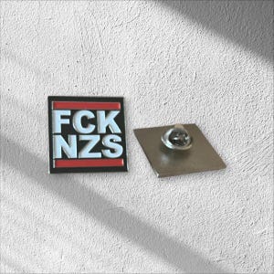 Könnte beinhalten: Quadratischer Emaille-Pin mit dem Text "FCK NZS" in weißen Blockbuchstaben auf schwarzem Hintergrund mit rotem Streifen. Ein silberfarbener, rechteckiger Pin-Rücken befindet sich neben dem Pin.