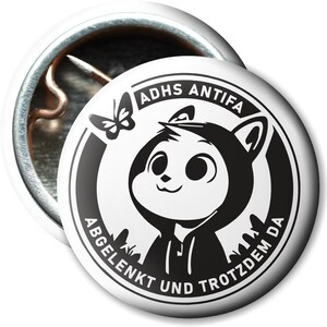 Könnte beinhalten: Schwarz-weißer Knopf mit einer Comicfigur, die eine Kapuzenjacke trägt und einen Schmetterling. Der Text auf dem Knopf lautet "ADHD Antifa Abgelenkt und trotzdem da".