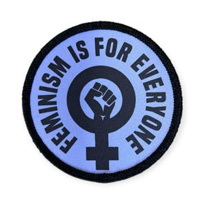 Könnte beinhalten: Runder Aufnäher mit dem Text "FEMINISM IS FOR EVERYONE" in schwarzer Schrift. Der Aufnäher hat einen hellblauen Hintergrund und einen schwarzen Rand. Ein schwarzes Symbol einer Faust in einem Kreis mit einem Kreuz darunter befindet sich in der Mitte.