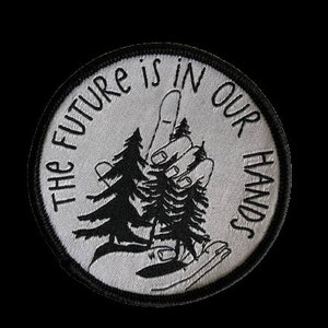 Könnte beinhalten: Ein runder, weißer gestickter Aufnäher mit schwarzen Nähten. Der Aufnäher zeigt eine Hand, die drei kleine Bäume hält. Der Text "The future is in our hands" ist um den Rand des Aufnähers herum geschrieben.