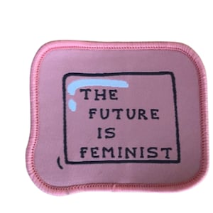 Könnte beinhalten: Rosa Stoff-Patch mit schwarzen Nähten und einem weißen Umriss. Der Patch trägt den Text "THE FUTURE IS FEMINIST" in einem schwarzen Rechteck.