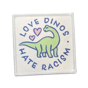 Könnte beinhalten: Quadratischer Aufnäher mit weißem Hintergrund und genähter Umrandung. Der Aufnäher zeigt einen grünen Dinosaurier mit blauen Umrissen, zwei rosa Herzen und den Text "LOVE DINOS. HATE RACISM" in Blau, alles in einer verspielten Schriftart.