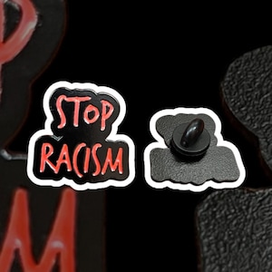 Könnte beinhalten: Ein schwarzer Emaille-Anstecker mit den Worten "STOP RACISM" in roter Schrift, weiß umrandet. Der Anstecker hat eine wolkenartige Form. Die Rückseite des Ansteckers hat einen schwarzen Stift und eine Kupplung.