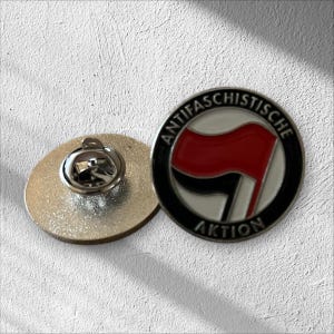 Könnte beinhalten: Runder Ansteck-Pin mit der Aufschrift "ANTIFASCHISTISCHE AKTION", die ein rotes Flaggen-Design auf schwarzem und weißem Hintergrund umgibt. Neben dem Pin befindet sich ein silberfarbener, runder Rücken.