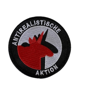 Könnte beinhalten: Ein schwarzer und weißer gestickter Aufnäher mit einer roten Einhorn-Silhouette. Der Text "Antirealistische Aktion" ist in Weiß um den Rand des Aufnähers geschrieben.