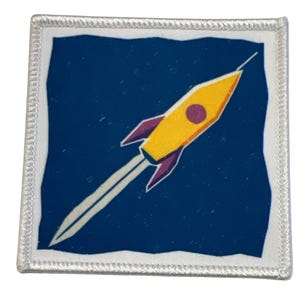 Könnte beinhalten: Ein weißer Patch mit blauem Hintergrund und einer gelben Rakete mit einer lila Flamme und einem lila Kreis an der Nase. Die Rakete fliegt nach oben.