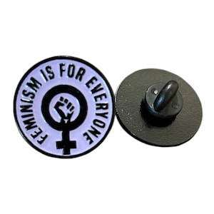 Könnte beinhalten: Runder Emaille-Anstecker mit dem Text "FEMINISM IS FOR EVERYONE" in weißen Buchstaben auf helllila Hintergrund. Der Anstecker zeigt eine schwarze Faust in einem weiblichen Symbol. Ein zweiter Anstecker mit schwarzer Rückseite ist ebenfalls sichtbar.