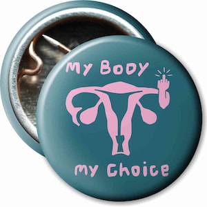 Könnte beinhalten: Ein blaugrüner Knopf mit einer rosa Gebärmutter und dem Text "My Body My Choice" mit einem erhobenen Mittelfinger.