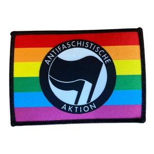 Könnte beinhalten: Ein rechteckiger Aufnäher mit Regenbogenhintergrund und schwarzem Rand. Der Aufnäher zeigt einen schwarzen Kreis mit einem weißen Flaggenmotiv und den Worten "ANTIFASCHISTISCHE AKTION" in einem kreisförmigen Muster.