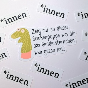Könnte beinhalten: Ein Aufkleber mit einer Cartoon-Sockenpuppe mit grün-gelbem Körper, rosa Manschette und großen Augen. Der Aufkleber hat Text auf Deutsch und ist von kleineren Aufklebern mit dem Wort "*innen" umgeben.