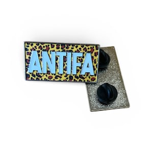 Könnte beinhalten: Ein rechteckiger Ansteckpin mit dem Wort "ANTIFA" in hellblauen Blockbuchstaben. Der Hintergrund hat ein Leopardenmuster in Braun- und Gelbtönen. Der Pin hat eine silberne Rückseite mit einem schwarzen Stiftverschluss.