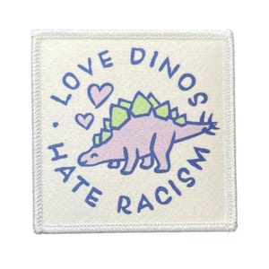 Könnte beinhalten: Quadratischer Aufnäher mit weißem Hintergrund und gesticktem Rand. Der Aufnäher zeigt einen lila Dinosaurier mit grünen Stacheln, zwei Herzen und den Text "LOVE DINOS HATE RACISM" in Blau.