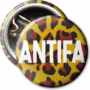 Könnte beinhalten: Zwei runde Ansteckbuttons mit einem Leopardenmuster in Gelb, Schwarz und Rot. Das Wort "ANTIFA" ist in weißen, fetten Buchstaben auf der Vorderseite aufgedruckt. Ein Button liegt leicht hinter dem anderen.