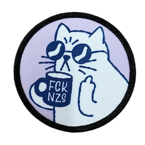 Könnte beinhalten: Runder Aufnäher mit einer weißen Katze mit Sonnenbrille, die eine Tasse mit dem Text "FCK NZS" hält und den Mittelfinger zeigt. Die Katze befindet sich auf einem hellvioletten Hintergrund mit schwarzem Rand.