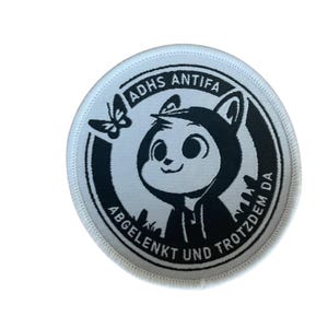 Könnte beinhalten: Ein weißer runder Patch mit schwarzem Text und einer Comicfigur. Der Text lautet "ADHS ANTIFA ABGELENKT UND TROTZDEM DA". Die Comicfigur ist ein schwarz-weißes Tier, das eine Kapuzenjacke trägt.
