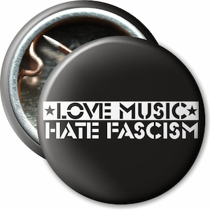 Könnte beinhalten: Ein schwarzer Button mit dem weißen Text "LOVE MUSIC HATE FASCISM" in Schablonenschrift. Der Button hat eine Metallrückseite und eine Anstecknadel. Das Design beinhaltet Sterne und eine kreisförmige Form.