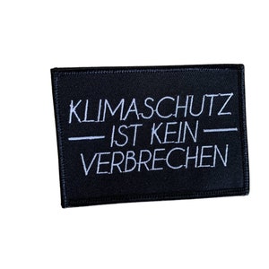 Könnte beinhalten: Schwarzer Stoff-Patch mit weißem Text, der "KLIMASCHUTZ IST KEIN VERBRECHEN" lautet.