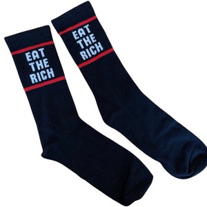 Könnte beinhalten: Ein Paar schwarze Socken mit einem roten Streifen und dem Text "Eat the Rich" in Weiß.