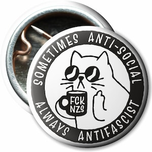 Könnte beinhalten: Ein weißer Button mit einer schwarz-weißen Illustration einer Katze mit Sonnenbrille und einer Tasse. Auf der Tasse steht "FCK NZS". Der Button trägt die Aufschrift "Sometimes Anti-Social Always Antifascist".