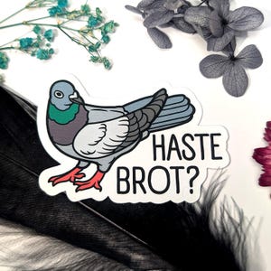 Könnte beinhalten: Ein Aufkleber mit einer Cartoon-Taube mit grauen, blauen und grünen Federn, roten Füßen und dem Text "HASTE BROT?" in Schwarz. Der Aufkleber befindet sich auf einem weißen Hintergrund mit dekorativen Elementen.