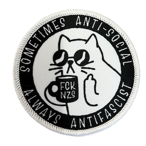 Könnte beinhalten: Runder, weißer Aufnäher mit schwarzem Rand. Er zeigt eine Cartoon-Katze mit Sonnenbrille, die einen Becher mit der Aufschrift "FCK NZS" hält und den Mittelfinger zeigt. Der Text am Rand lautet "SOMETIMES ANTI-SOCIAL ALWAYS ANTI-FASCIST".